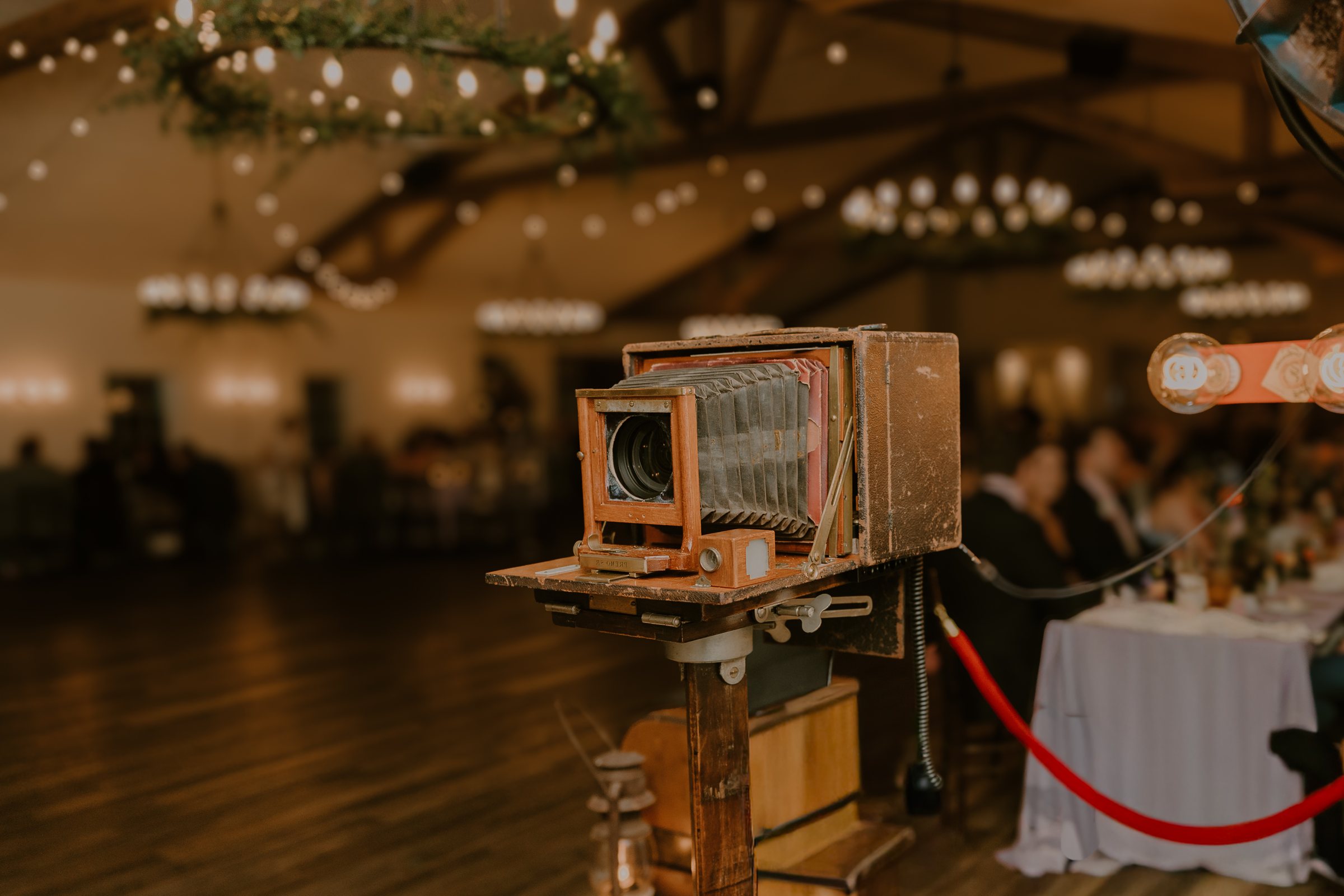 A vintage bellows camera on its stand at a warmly lit wedding reception, string lights glowing overhead.